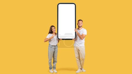 Téléchargez les photos : A young woman and man happily display a large vertical smartphone, pointing at its blank screen for potential advertisement on a goldenrod background. Attention, good news, website - en image libre de droit