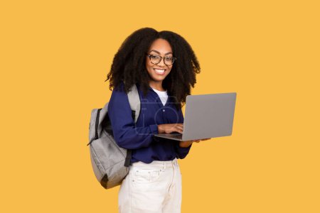Photo for Cheerful young black lady with backpack and headphones holding laptop and smiling at camera over yellow background. Distance learning and remote education concept - Royalty Free Image