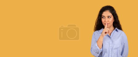 Téléchargez les photos : Curious arab young woman with long curly hair placing her finger on lips in a shushing gesture, looking to the side with a secretive expression on a yellow background, studio - en image libre de droit