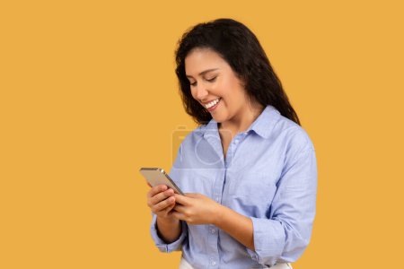 Téléchargez les photos : A cheerful young woman with long black hair, wearing a light blue shirt, is happily looking at her smartphone on a vibrant orange background. App for work online recommendation - en image libre de droit