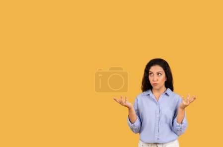 Photo for Puzzled pensive arab young woman in a light blue shirt with raised hands, showing a questioning gesture, standing against a plain yellow background with a look of uncertainty - Royalty Free Image