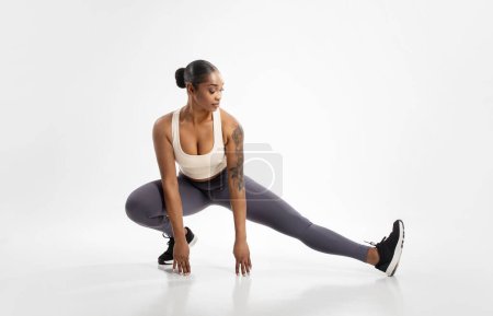 Photo for Fit black lady making extended leg squat pose stretching legs, exercising against white background. Full length studio shot of athlete woman having workout for muscles flexibility - Royalty Free Image