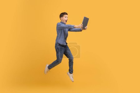 Foto de Surprised teenage boy jumping with digital tablet in hands over yellow background, happy teen kid showing excitement or shock, enjoying modern technologies, full length with copy space - Imagen libre de derechos