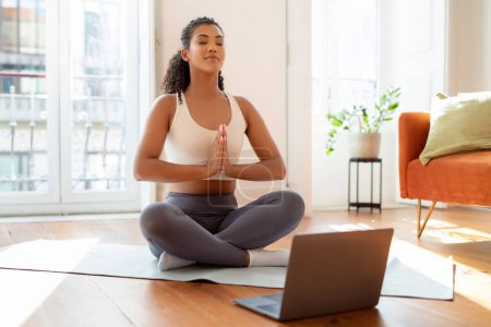 Foto de Fit young lady practicing yoga and meditation in front of her laptop indoors, sitting on mat in lotus position, meditating for mindfulness and balance at modern home interior - Imagen libre de derechos