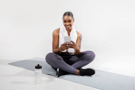 Téléchargez les photos : Black fitness woman browsing online workout on smartphone, taking break from training on mat, sitting with bottle of water and cellphone over white studio background. Sport applications - en image libre de droit