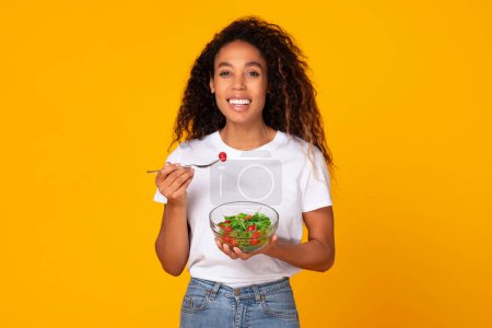 Photo for African American woman happily eating a nutritious vegetable dish, illustrating diet lifestyle on yellow background. Slim lady holds salad bowl savoring vegetables on dinner - Royalty Free Image