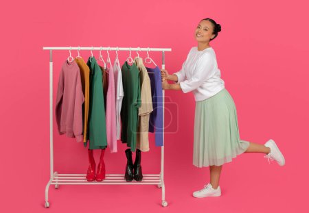 Foto de Happy Asian woman standing near clothes rack, showcasing variety of outfits, happy shopaholic lady demonstrating new trendy collection, posing on pink studio background, copy space - Imagen libre de derechos