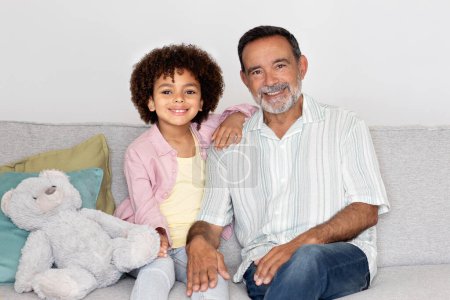 Téléchargez les photos : Cheerful Brazilian grandfather and grandson posing sitting together on a couch indoors, showcasing joyful family moment in warm home setting, embracing with bright smiles - en image libre de droit