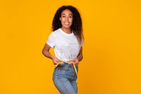 Foto de Black young lady in jeans holds measuring tape around her slim waist, posing on yellow studio backdrop, smiling to camera, happy about successful slimming. Woman measuring weight loss result - Imagen libre de derechos