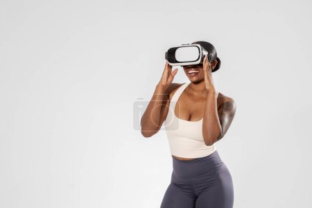 Téléchargez les photos : Young black woman in activewear immersed in VR exercise, using virtual reality headset for sport training or gaming, standing on white backdrop, showcasing futuristic fitness workout. Free space - en image libre de droit
