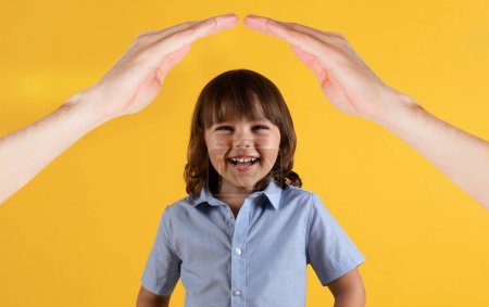Photo for Child safety concept, insurance for kids. Cute laughing blonde boy preschooler child in blue shirt standing under woman hands roof, yellow studio background - Royalty Free Image