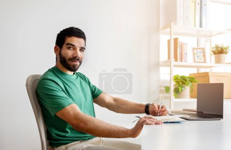 Photo for Smiling bearded man with glasses in a green shirt sitting at a desk in a bright office, writing in a notebook with a laptop open in front of him, shelves with books and plants in the background - Royalty Free Image