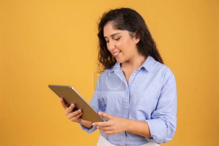 Photo for Engrossed young woman with curly hair in a light blue shirt reads content on a digital tablet, with a warm yellow background enhancing her focus. Study, work online recommendation - Royalty Free Image