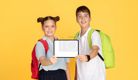 Foto de A schoolgirl and schoolboy with backpacks smilingly present a tablet with a white screen, ready for customization, against a yellow background, suggesting interactive learning - Imagen libre de derechos