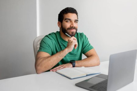 Téléchargez les photos : Confident middle eastern man with a beard and a green t-shirt sitting at a desk with a laptop and notebook, pen in hand, looking at the camera, in a clean and bright office - en image libre de droit