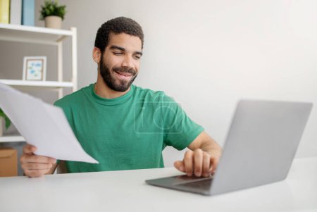 Photo for Smiling young middle eastern man in green t-shirt happily reviewing documents while simultaneously working on a laptop in a home office setting, showcasing multitasking at home - Royalty Free Image