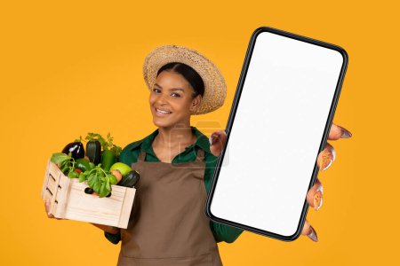 Téléchargez les photos : Smiling Black Lady Farmer Holding Wooden Box With Fresh Farm Vegetables And Showing Smartphone With White Blank Screen, Posing Over Yellow Studio Background. Online Business, E-commerce - en image libre de droit