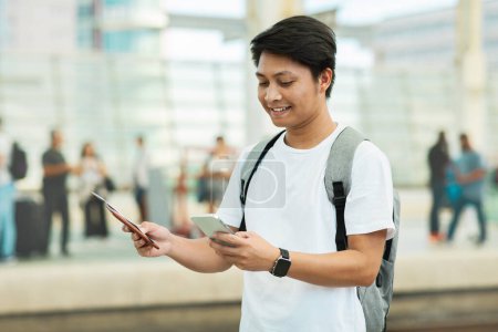 Photo for Portrait Of Young Asian Man Using Smartphone While Waiting At Airport, Happy Smiling Chinese Guy Checkin Online On Mobile Phone While Standing With Backpack At Crowded Terminal, Copy Space - Royalty Free Image