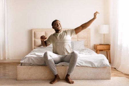 Téléchargez les photos : Good Morning. Happy Black Man Waking Up And Stretching Arms With Toothy Smile, Relaxed After Great Rest, Sitting In Bed In Modern Bedroom Interior. Start Of New Day, Rest And Wellness Concept - en image libre de droit