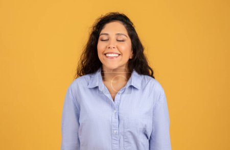 Téléchargez les photos : Radiant young woman with closed eyes and a joyful smile, wearing a light blue shirt, exudes happiness and contentment on a mustard yellow background. Freedom, positive lifestyle, relax - en image libre de droit