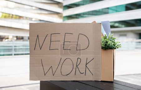 Téléchargez les photos : Cardboard sign with Need Work text alongside personal items standing outdoors against modern business center on background, conceptual image for unemployment and joblessness problem - en image libre de droit