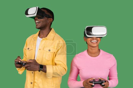 Foto de Excited african american young man and woman using VR glasses and consoles joysticks, black couple enjoy play game in virtual reality, isolated on green background, studio. Fun with technology - Imagen libre de derechos