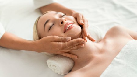 Foto de Young Blonde Woman Enjoying Relaxing Massage Lying On Bed, Masseur Massaging Her Neck And Face At Spa Indoors, Closeup. Relaxation, Beauty And Wellness Concept. Panorama - Imagen libre de derechos