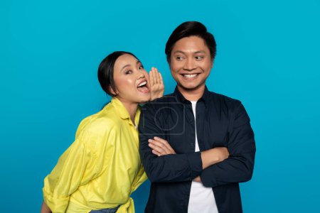 Photo for An Asian millennial woman whispers into the ear of a smiling man with arms crossed, sharing a secret or making a playful comment, against a bright turquoise backdrop, studio - Royalty Free Image