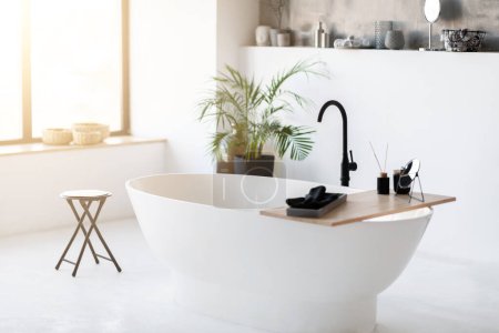 Téléchargez les photos : Spacious, modern bathroom interior featuring sleek freestanding bathtub in the middle, room filled with sunlight with minimalist luxury decor and potted plant in corner, copy space - en image libre de droit