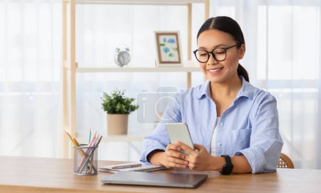 Foto de Pretty smiling young asian woman wearing eyeglasses sitting at desk with laptop, using smartphone, chinese lady entrepreneur using business mobile app, working from home, copy space - Imagen libre de derechos