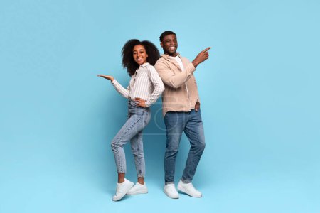 Photo for Cheerful young black man and woman standing back to back, smiling and pointing to their sides at free space against serene blue background - Royalty Free Image