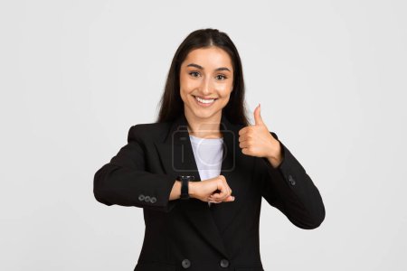 Foto de Happy professional woman in suit giving thumbs up while looking at her smartwatch, representing efficient time management, against light grey background - Imagen libre de derechos