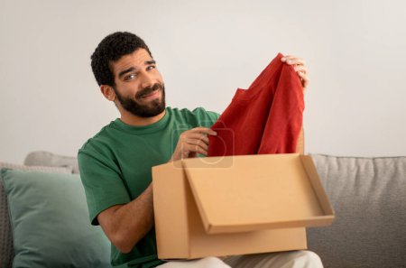 Téléchargez les photos : Smiling middle eastern young man in a green shirt skeptically examines a red sweater just unpacked from a cardboard box, seated on a grey couch in a cozy living room at home - en image libre de droit