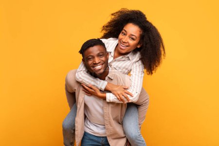Téléchargez les photos : Cheerful young black man giving a piggyback ride to an excited woman, both sharing genuine smile, set against warm yellow background radiating happiness - en image libre de droit