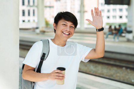 Foto de Positive young asian guy tourist waving and smiling at camera, holding takeaway coffee cup, carrying backpack, waiting for train at station. Chinese man meeting friend or lover at railway stop - Imagen libre de derechos
