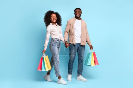 Téléchargez les photos : Happy young black couple holding hands and carrying colorful shopping bags, walking with relaxed, content smiles on vibrant blue background - en image libre de droit