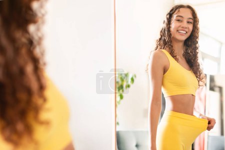 Téléchargez les photos : Happy fit teen girl looking at reflection in mirror and pulling fitness pants waist showing result of dieting and slimming, motivated after workout training at home. Sport and weight loss - en image libre de droit