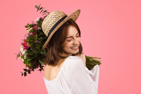 Photo for Joyful happy european woman in a summer outfit, with a straw hat and sunglasses, holding a bouquet of fresh flowers, enjoying the moment on a vibrant pink studio background - Royalty Free Image