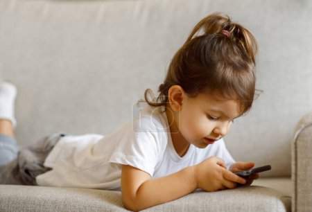 Téléchargez les photos : Cute Little Girl Playing Online Games On Smartphone While Lying On Couch, Adorable Preschool Female Child Using Mobile Phone For Leisure, Relaxing On Sofa In Living Room At Home, Closeup - en image libre de droit