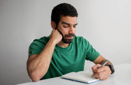Téléchargez les photos : Serious man with a beard in a green t-shirt writing in a notebook, resting his chin on his hand, looking focused at his work, at a white desk with a smartwatch on his wrist - en image libre de droit