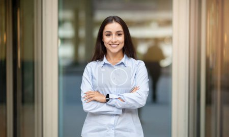 Téléchargez les photos : A confident happy pretty caucasian young woman manager with long hair, standing with crossed arms, wearing a light blue shirt and a smartwatch, smiling in an office corridor - en image libre de droit