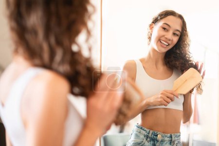 Téléchargez les photos : Happy Teen Girl Brushing Hair With Wooden Hairbrush, Smiling To Her Reflection In Mirror At Home, Selective Focus. Young Lady Enjoying Her Daily Haircare Routine In The Morning - en image libre de droit
