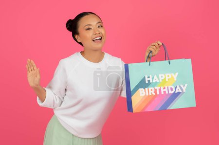 Happy young asian woman holding paper bag with Happy Birthday text, cheerful korean female preparing for celebration, enjoying gifts, standing isolated on pink studio background, copy space
