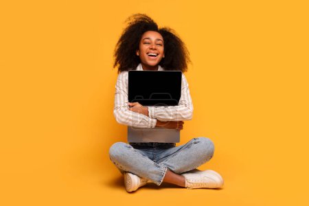 Photo for Joyful young black woman with curly hair, embracing blank screen laptop, sitting cross-legged against vibrant yellow background, exuding happiness - Royalty Free Image