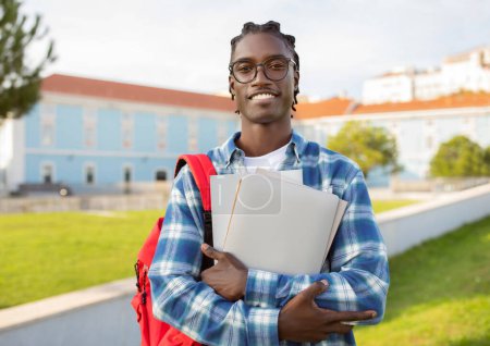 Foto de Cheerful black college student guy stands with his laptop, backpack and books smiling to camera at university park outside, wearing glasses. Education and academic success concept - Imagen libre de derechos