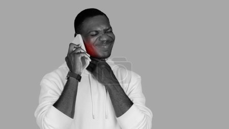 Foto de A monochrome image shows african american man in a white hoodie wincing in pain with a red mark on his cheek while talking on the phone, adding a splash of color to the grayscale scene - Imagen libre de derechos