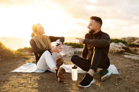 Téléchargez les photos : Man and woman sharing joyful moment while resting post workout, seated on mat with water bottles, against stunning sea backdrop at sunset - en image libre de droit