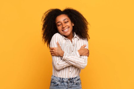 Foto de Young black woman with curly hair hugging herself, eyes closed with joyful smile, exuding self-love and happiness on warm yellow background - Imagen libre de derechos