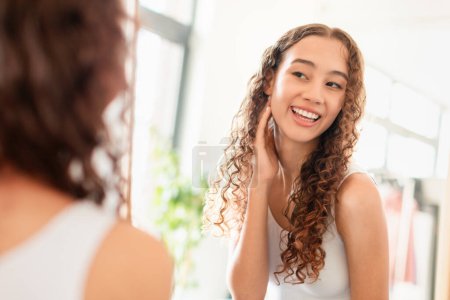 Téléchargez les photos : Happy Teen Girl Touching Smooth Face Looking In Mirror At Home, Young Lady Looks At Her Reflection Enjoying Fresh Perfect Skin In The Morning. Youth Skincare Routine And Cosmetics Advertisement - en image libre de droit