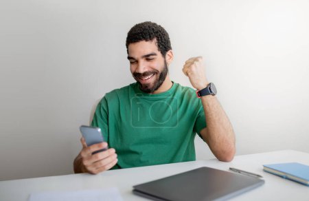 Photo for Joyful man middle eastern with a beard in a green t-shirt celebrates good news on his smartphone at his white work desk, with a smartwatch on his wrist and a laptop nearby - Royalty Free Image
